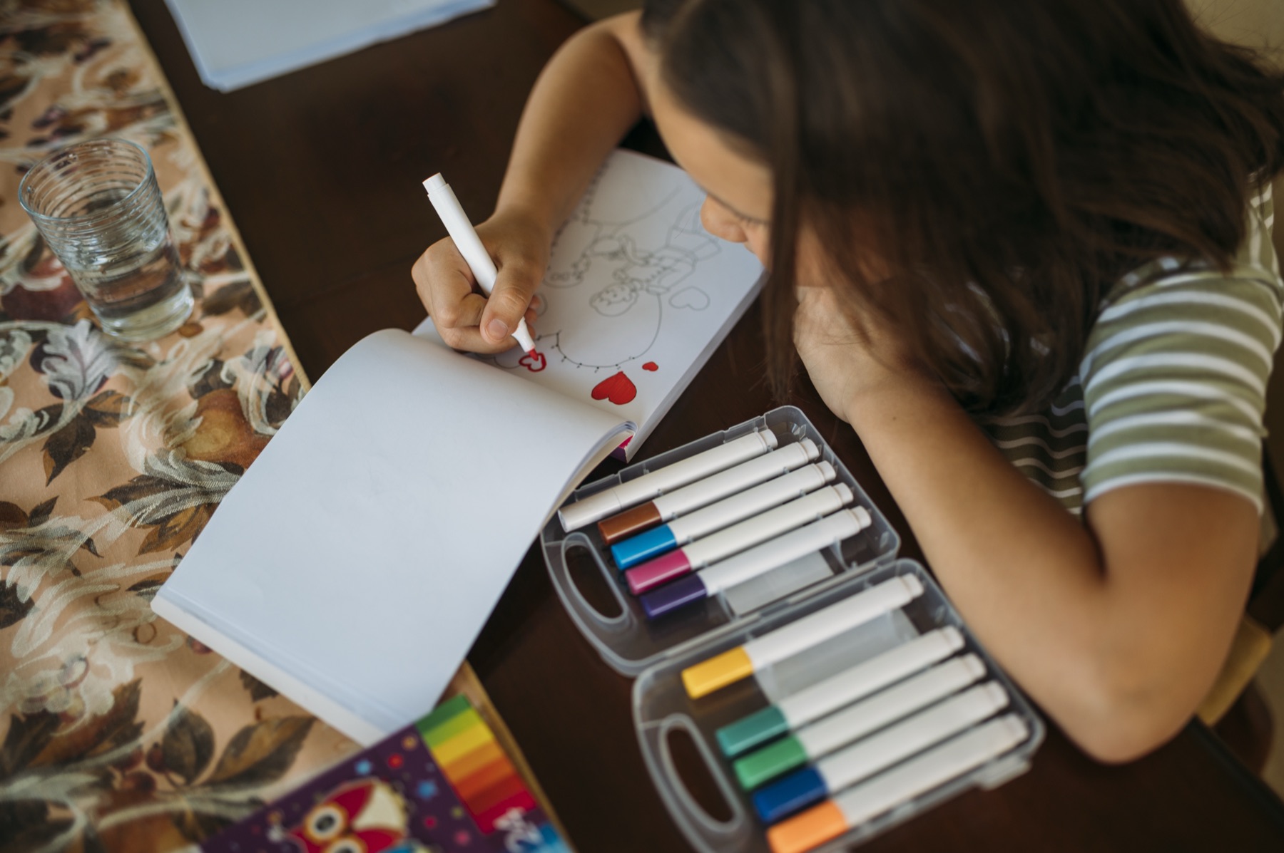 enfant qui colorie un dessin déjà tracé avec des feutres lavables rangés dans une boîte à côté d'un verre d'eau